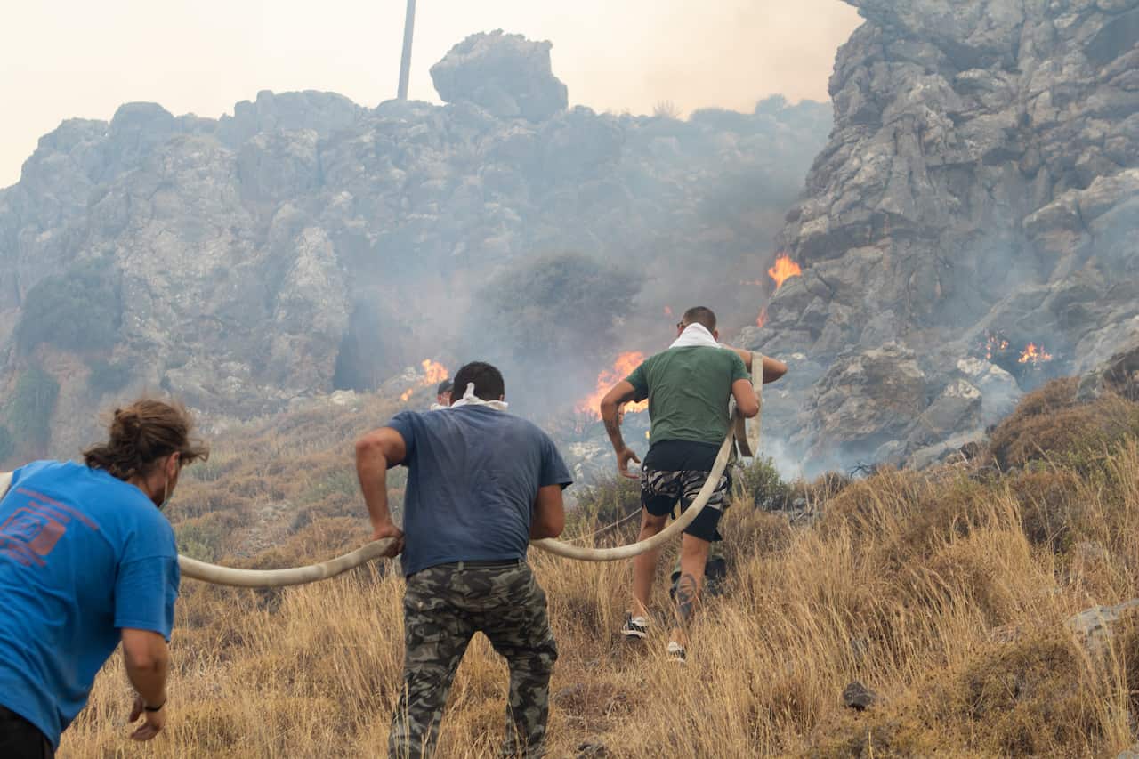 Four firefighters on a mountain carry a long hose towards fires in the distance.