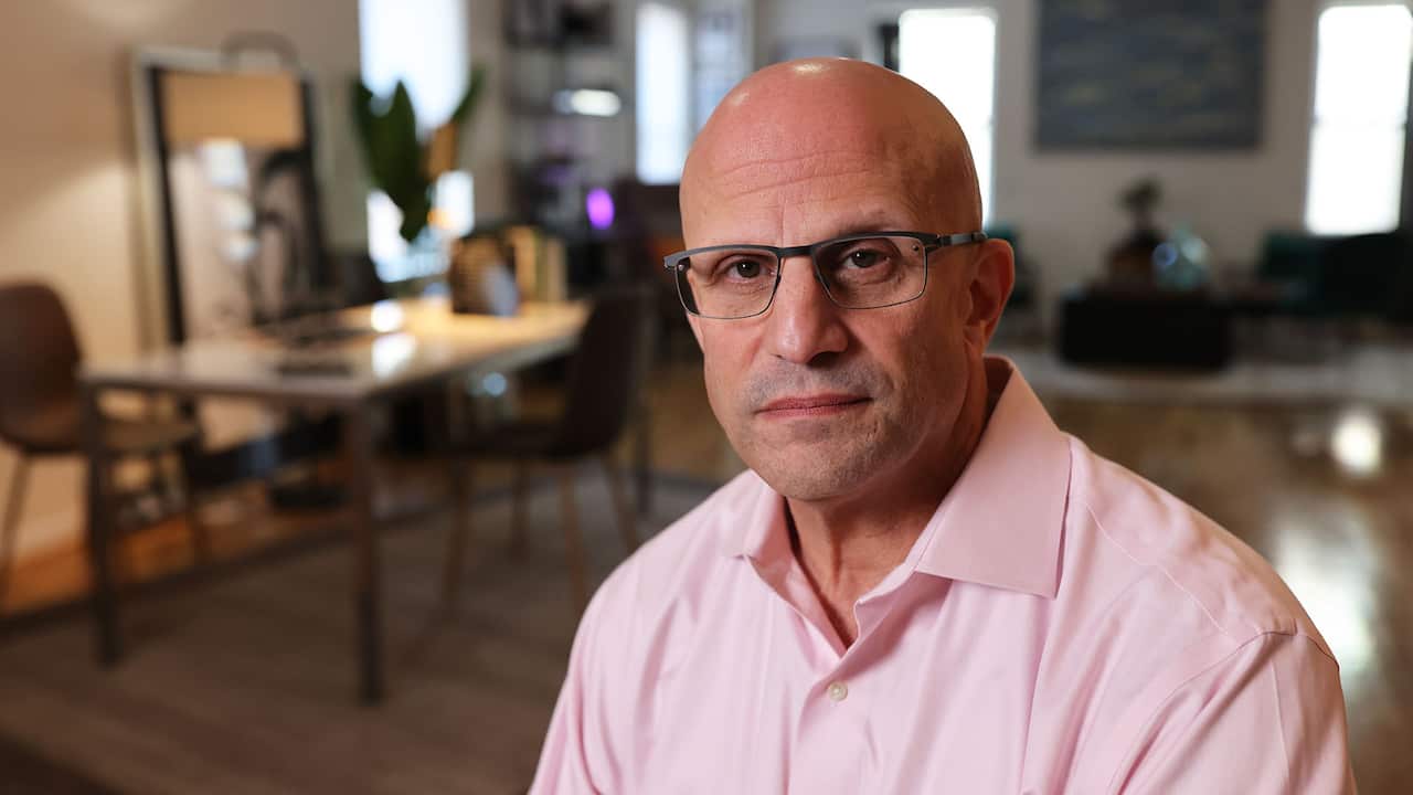 A bald middle-aged man wearing reading glasses and a light pink shirt 