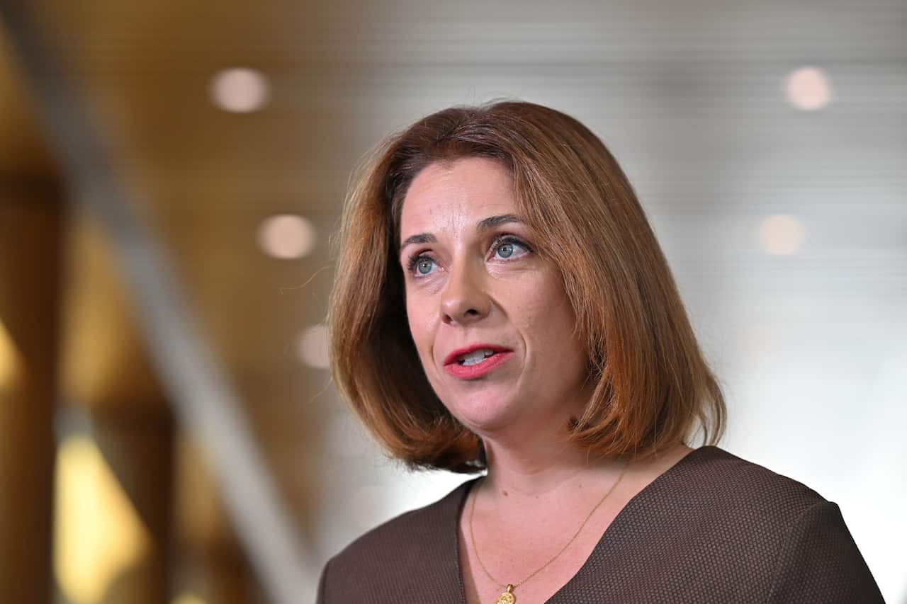 A medium shot shows a woman with brown hair and a dark top looking slightly up and to the left. She has a subtle smile and appears to be speaking.