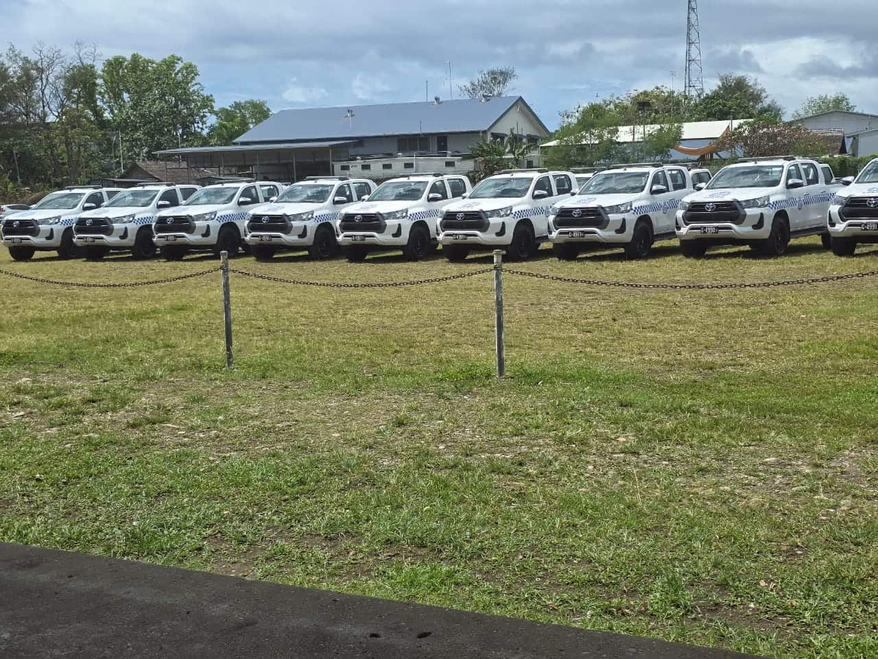 A fleet of white SUVs is parked in an open green space with a fence in the foreground.