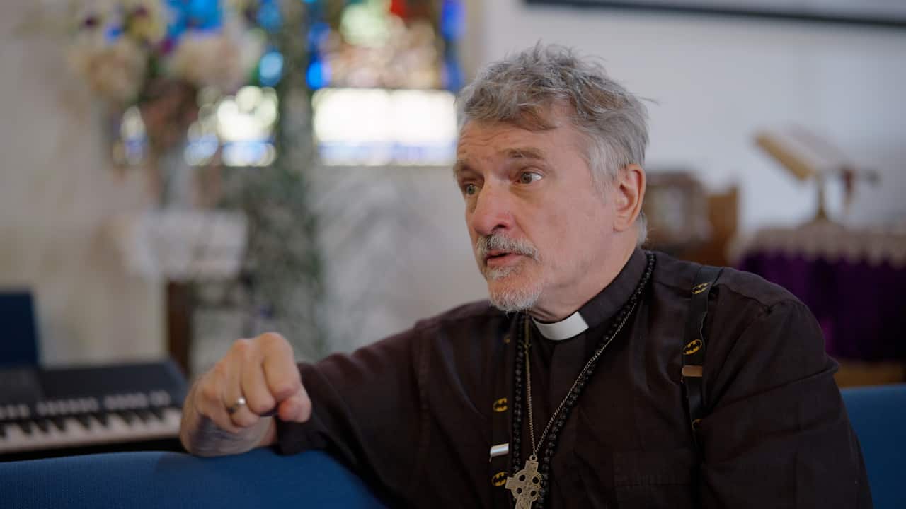 Priest in black clothing and white dog collar giving an interview.