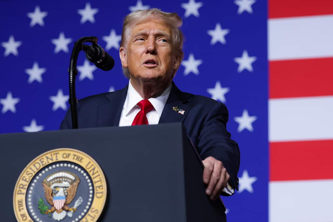 A man in a dark suit with a red tie standing at a podium in front of the American flag