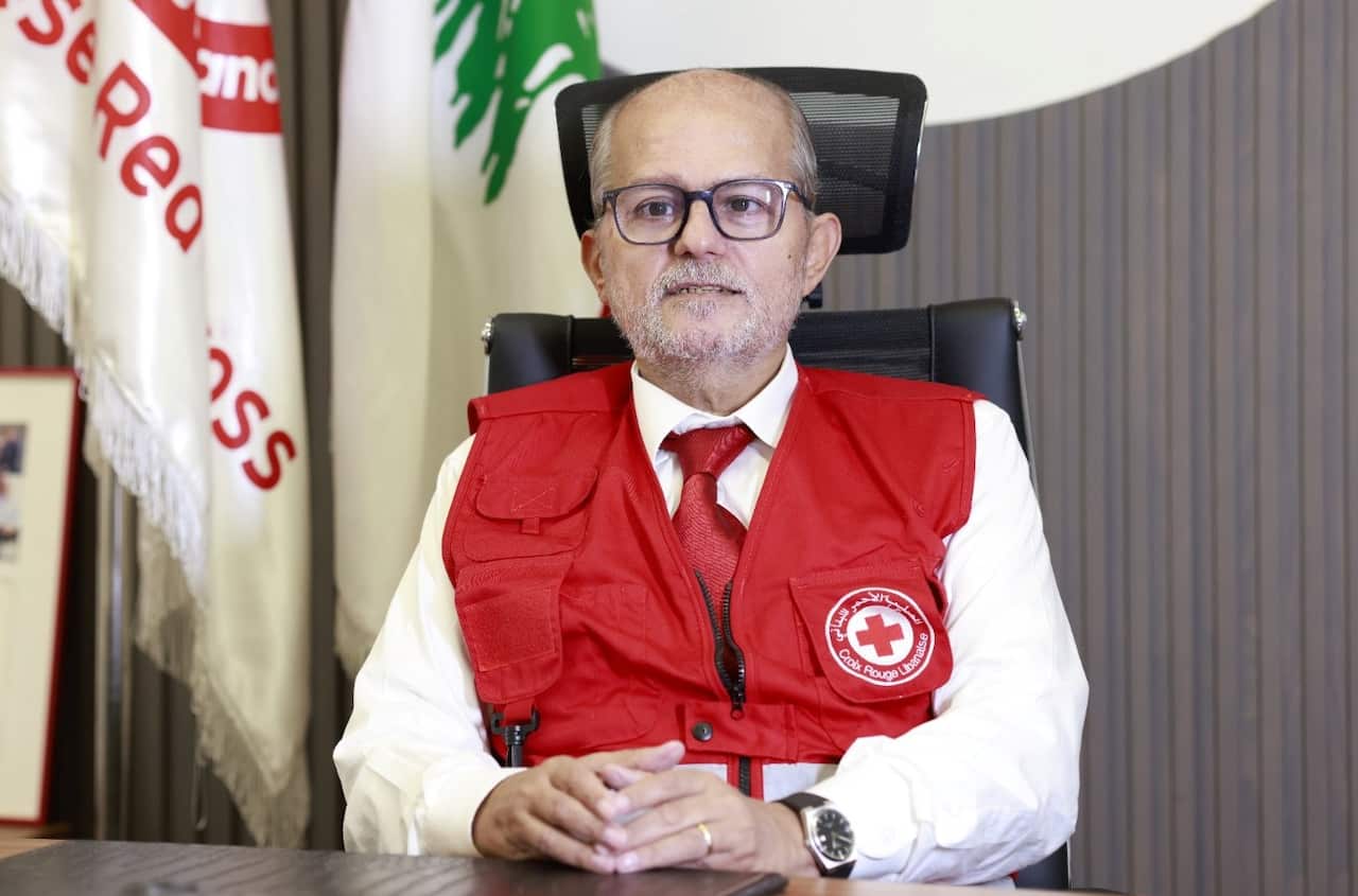 Georges Kettaneh, sits at a desk wearing glasses and a red vest with a Red Cross emblem