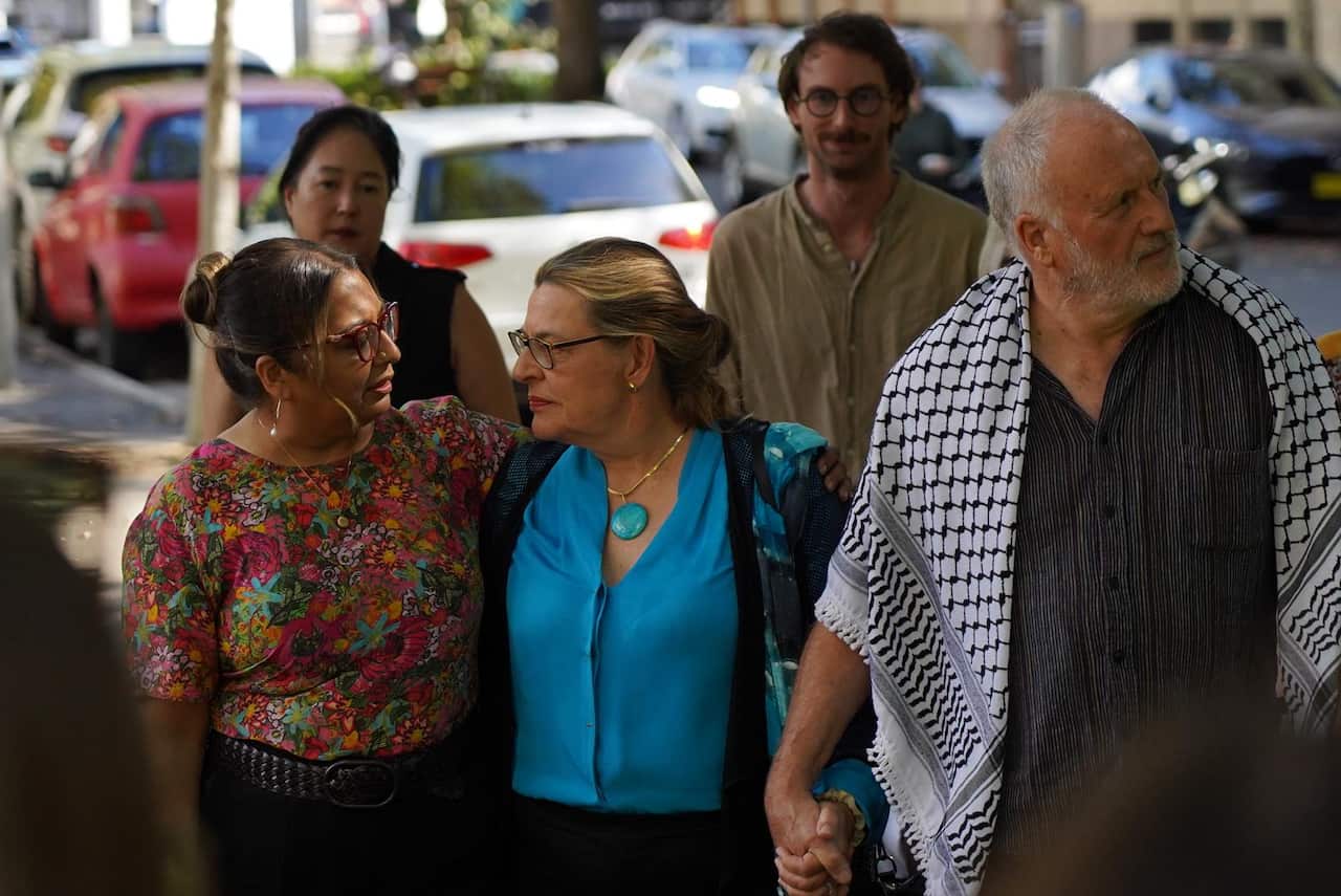 A distressed-looking middle-aged woman is comforted by another woman while a taller man with white hair beside them looks to the right.