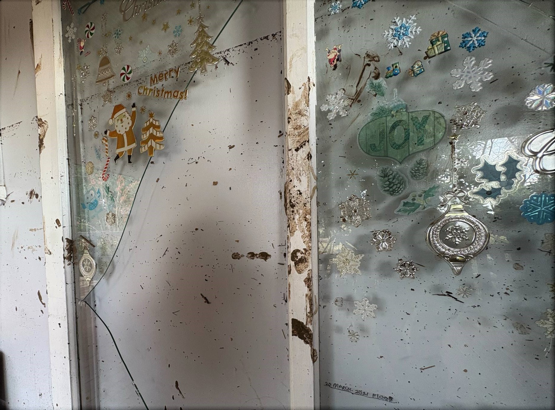 A broken glass door that has Christmas decorations on it. On the white wall on the bottom of the frame is a marking that reads 'March 2021 flood'. Above it is a line on the wall showing the 2025 flood level far surpassing 2021.