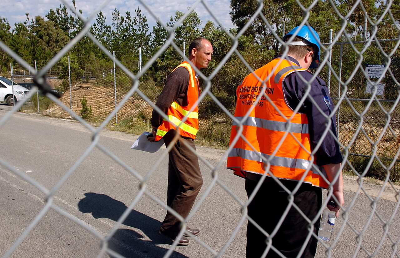 Two men in high viz gear stand behind a wire fence.