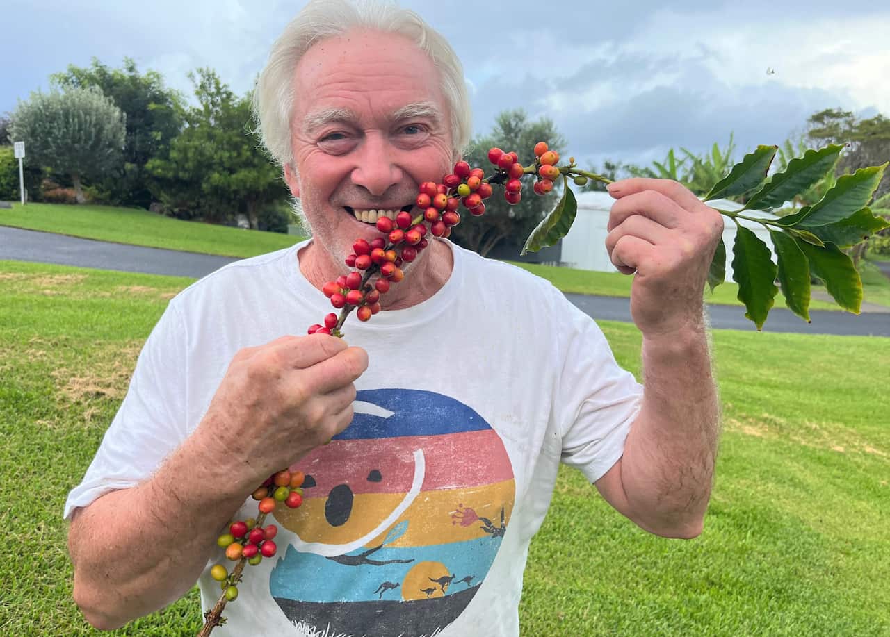 A man in a white t-shirt stands holding a branch covered in red berries.