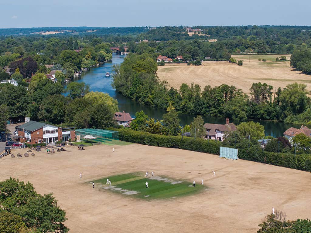 Aerial view of a cricket field in England that is largely light brown because of drought. There are trees and a river in the distance