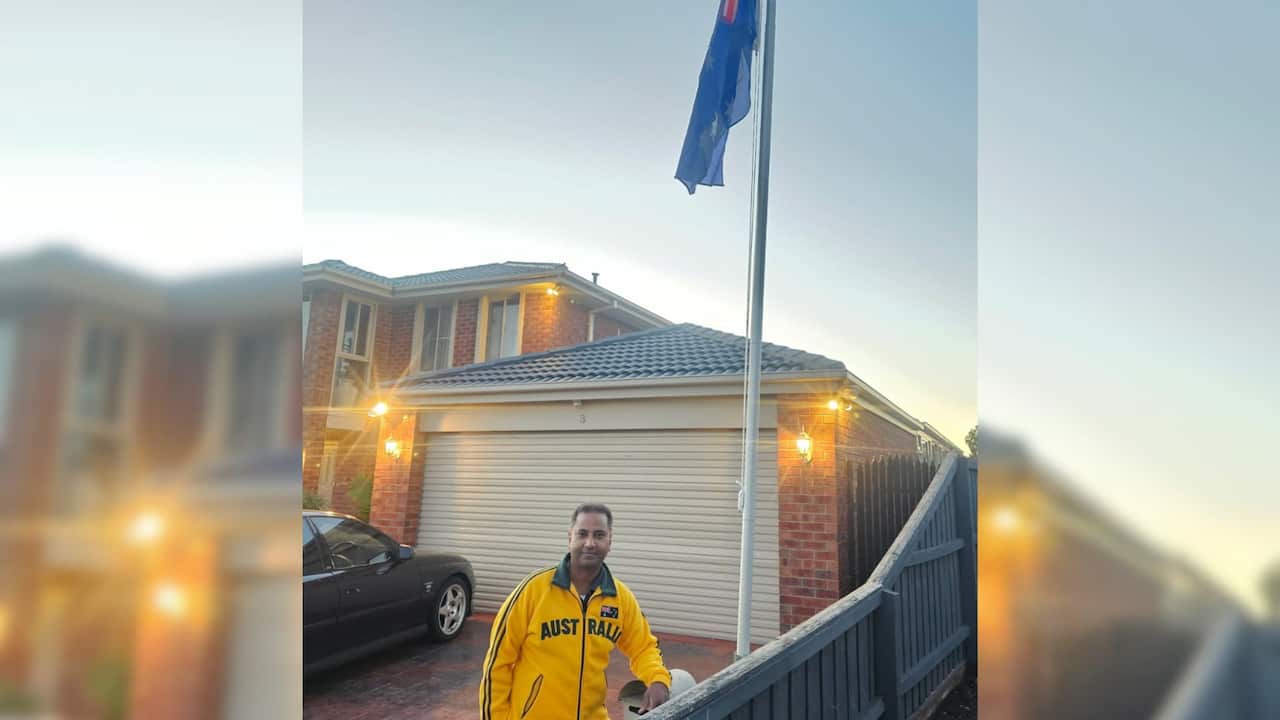 a man in an Australian green and gold jacket stands on a driveway under a flapole with an Australian flag