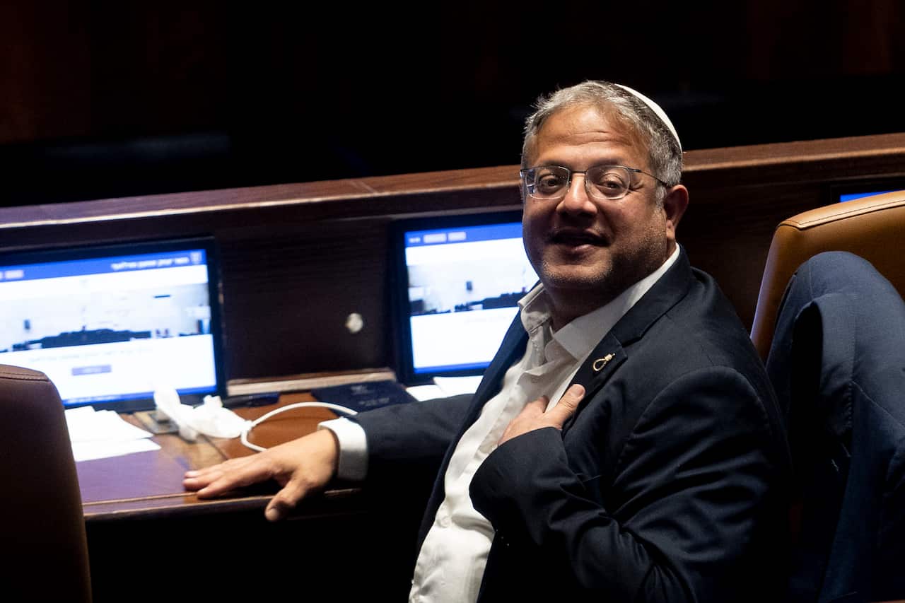 An Israeli man with glasses and a white kippah smiles while seated at a dark wooden desk equipped with computer monitors, holding one hand to his chest.