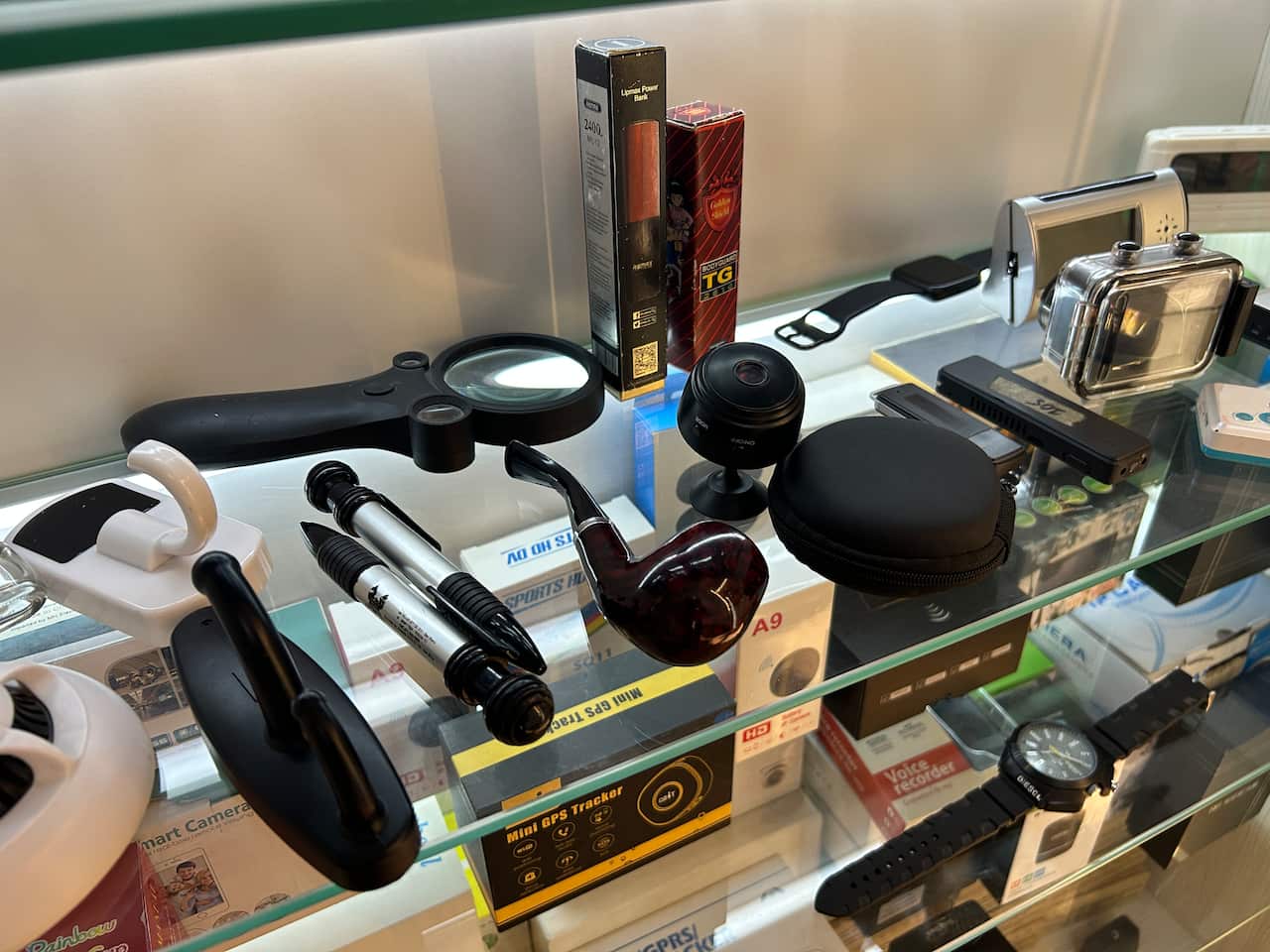 A collection of gadgets on a glass shelf, including pens, a pipe and a magnifying glass. Below is another shelf with cameras, a mini GPS trackers, and assorted boxes. 