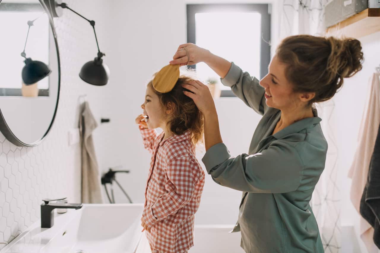 Mother with small child indoors in bathroom in the morning at home, brushing teeth and hair.
