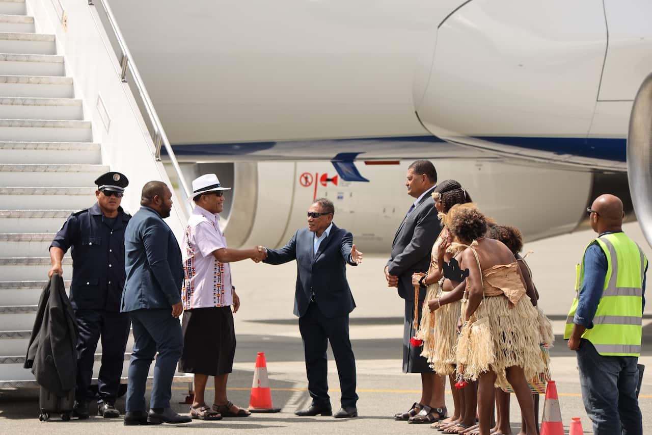 A group of men in suits and traditional dress are gathered at the foot of an airplane staircase on an airport tarmac. A man in a suit is shaking hands with a man wearing a white hat, while a group of people in traditional woven skirts stand to the side.