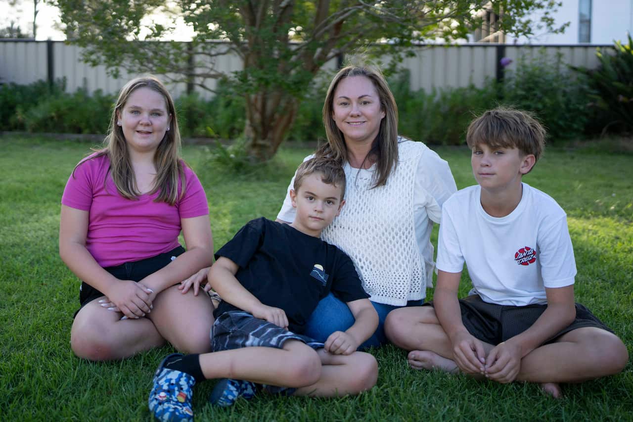 A middle-aged woman sits in a grassy backyard surrounded by three children: two boys and a girl.