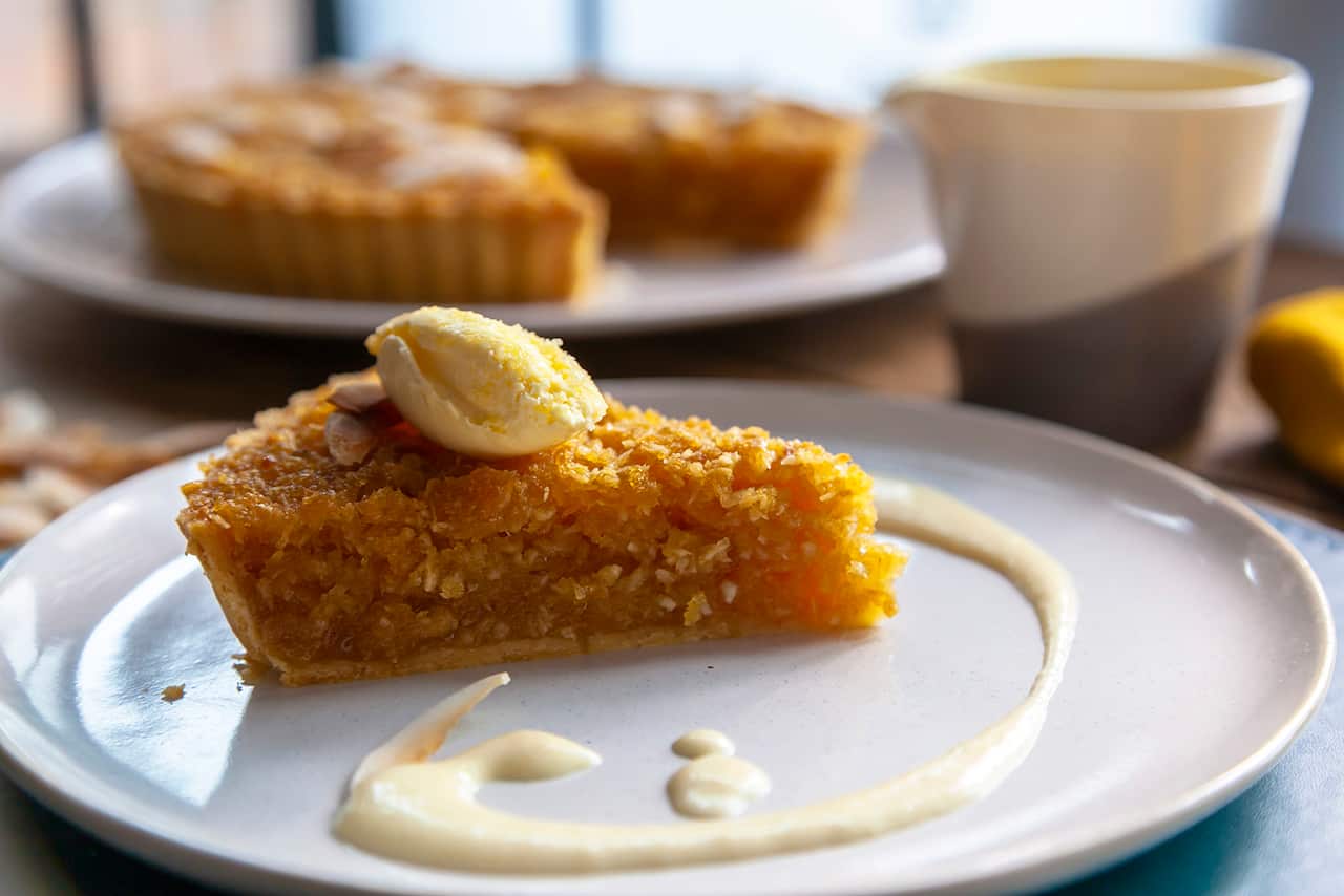 A wedge-shaped slice of orange-gold tart sits on a low, pale round plate, with a swirl of cream. The rest of the tart can be seen in the background. 