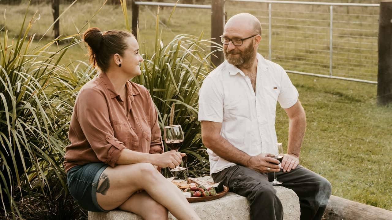 a woman and a man in casual clothes sitting on a block sharing a cheese board in front of a green paddock
