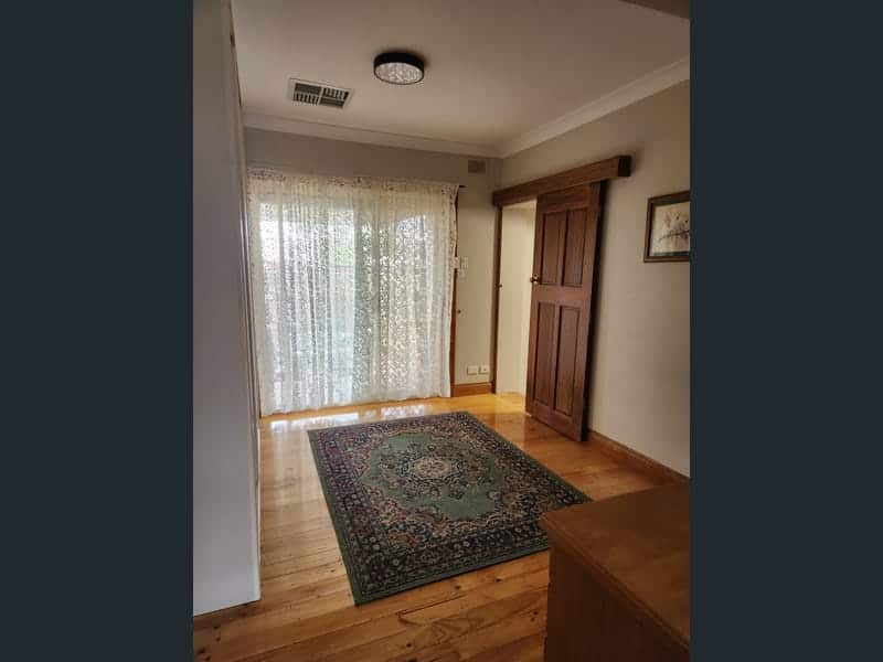 The interior of a house looking toward a window with a white lace curtain, an oriental rug on the floor, and polished wooden flooring.