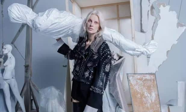 A model with blonde hair wearing a studded leather jacket and holding a mannequin wrapped in a white sheet