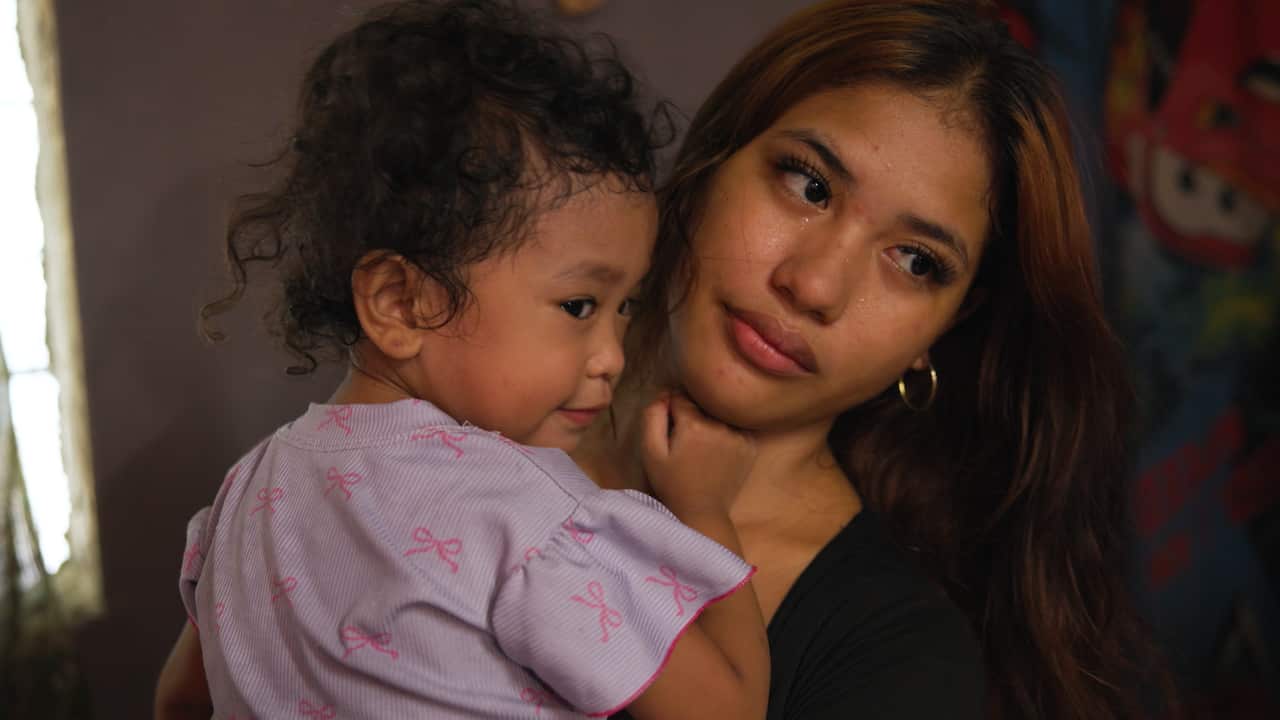 A young Filipino woman with long hair holds a small child wearing a pink dress with a bow pattern. The woman has tears rolling down her cheeks.