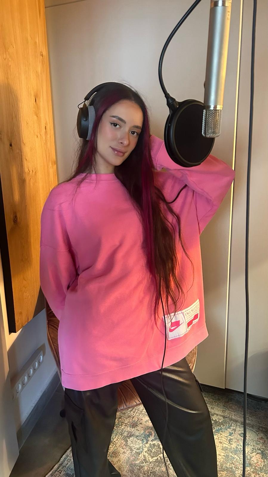 A musician in a pink top wearing headphones stands in front of a microphone.