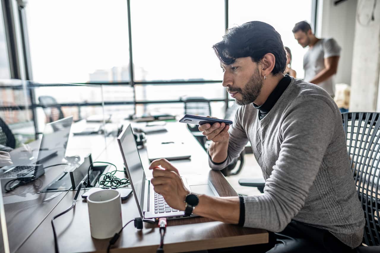 A man working at a hot-desk on his laptop with his mobile phone towards his mouth.