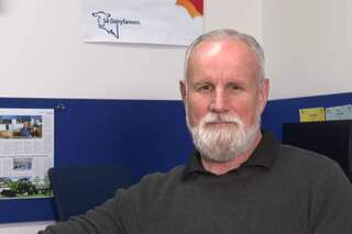 John Elferink, wearing a grey jumper, sitting in front of a blue cubicle and white wall.