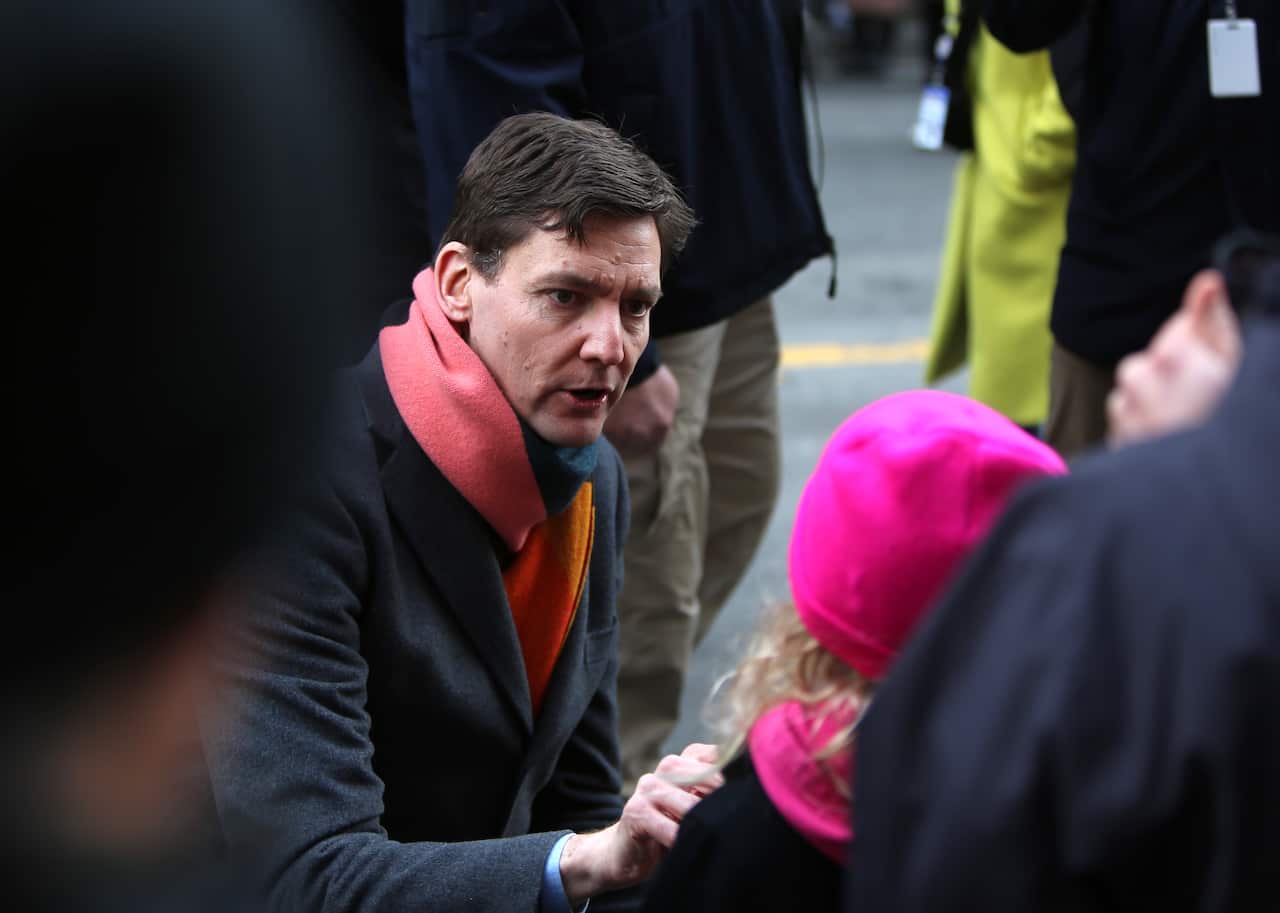 A man wearing a dark winter coat and a red scarf is squatting in front of a girl in a pink hat 