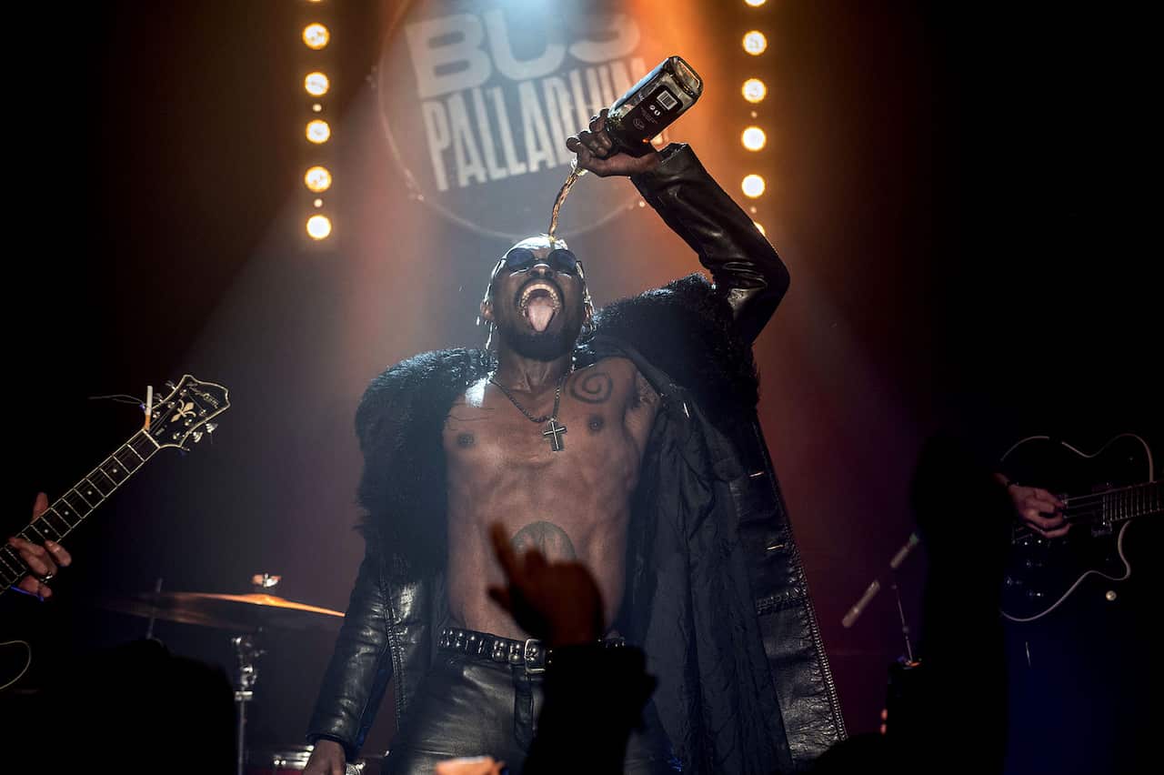 A bare-chested man in a long black coat and black leather pants stands on stage, pouring alcohol from a bottle on his head. Drums and a guitar can be seen behind him.