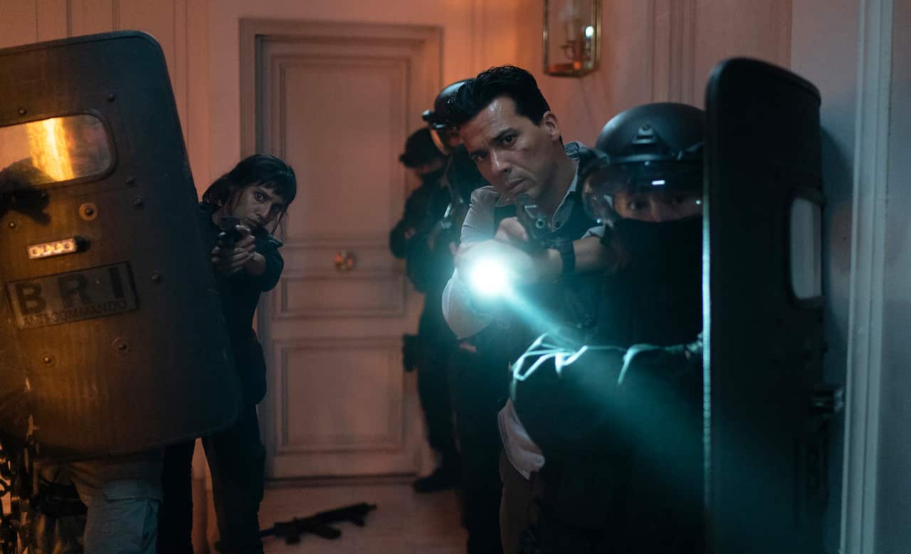 In the midst of a group of riot police, a man stands holding a gun, peering ahead. A woman holding a gun stands in the group, to the rear.  