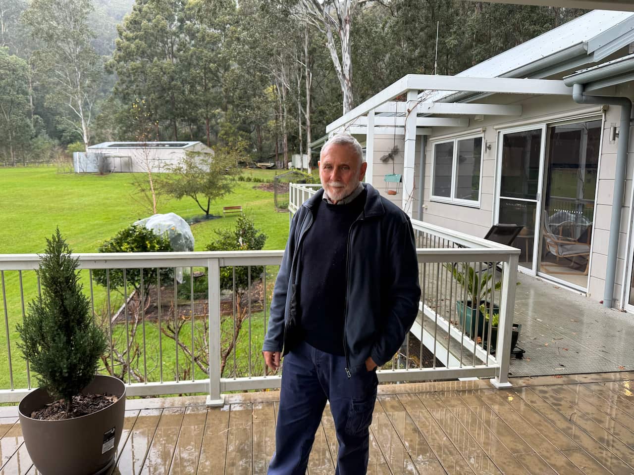 A man stands on his porch overlooking a garden with water tank in the background