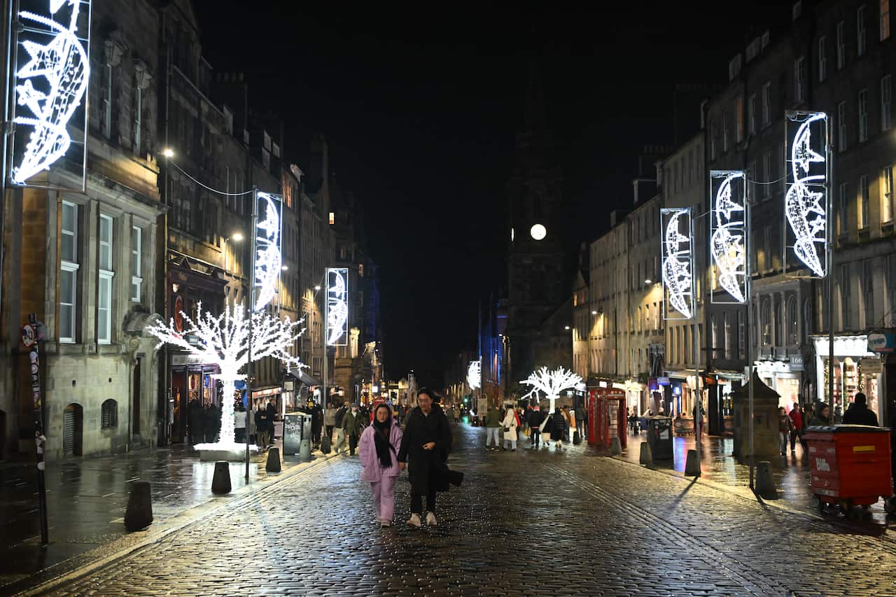 People stroll along a lit cobbled street flanked by medieval buildings.
