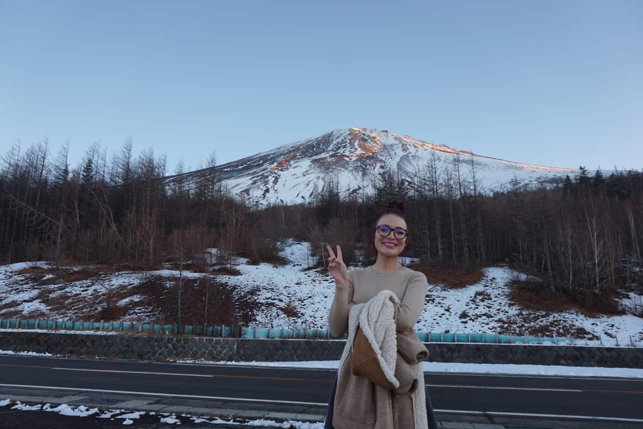 A woman holds up a peace-sign and smiles in front of a round and a snowy mountain. 