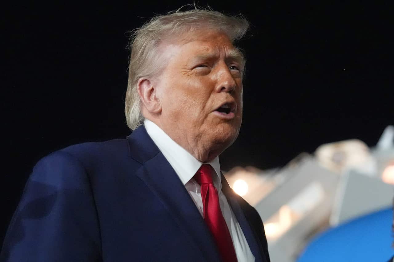Close-up side profile of an older man with light blond hair wearing a dark navy suit, white shirt, and red tie, speaking with his mouth slightly open. The background is out of focus with bright lights and a large aircraft-like structure, suggesting an outdoor nighttime event.