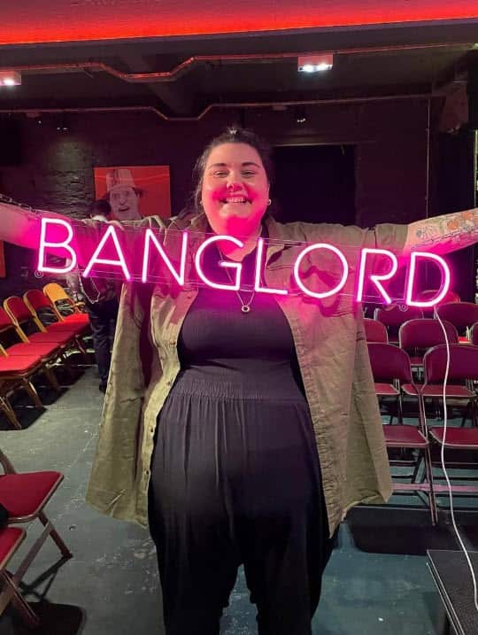 A person smiling while standing in an underground performance venue and holding a fluorescent pink sign that says "banglord".