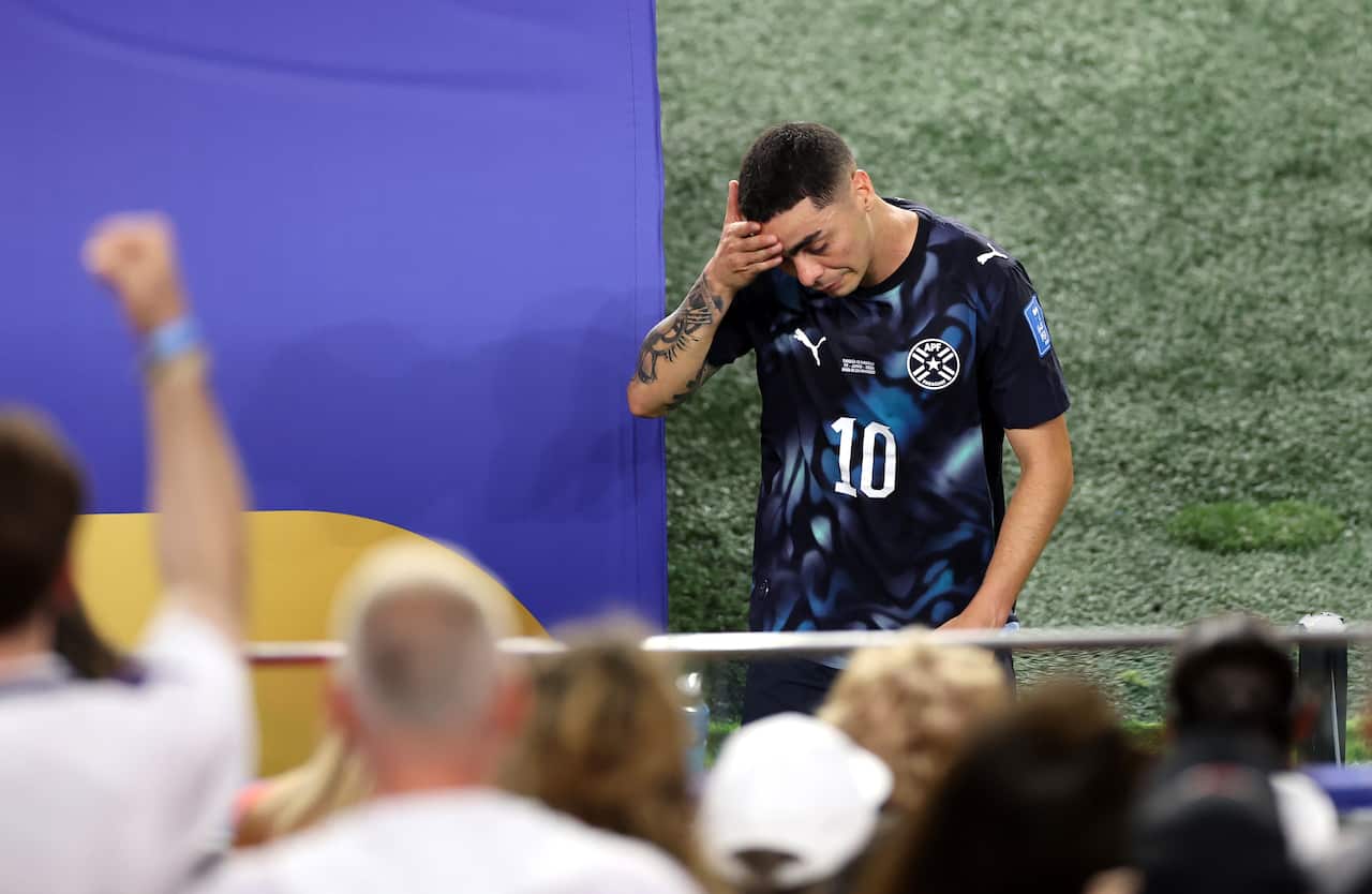 Paraguay player wearing the number 10 jersey touches his forehead as he walks past a barrier near the pitch, with out-of-focus spectators in the foreground.