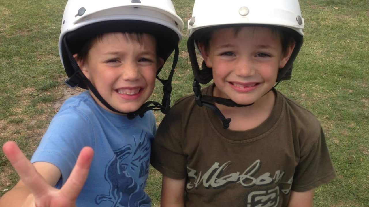 two young boys with white helmets on stand on the grass smiling and giving the peace sign