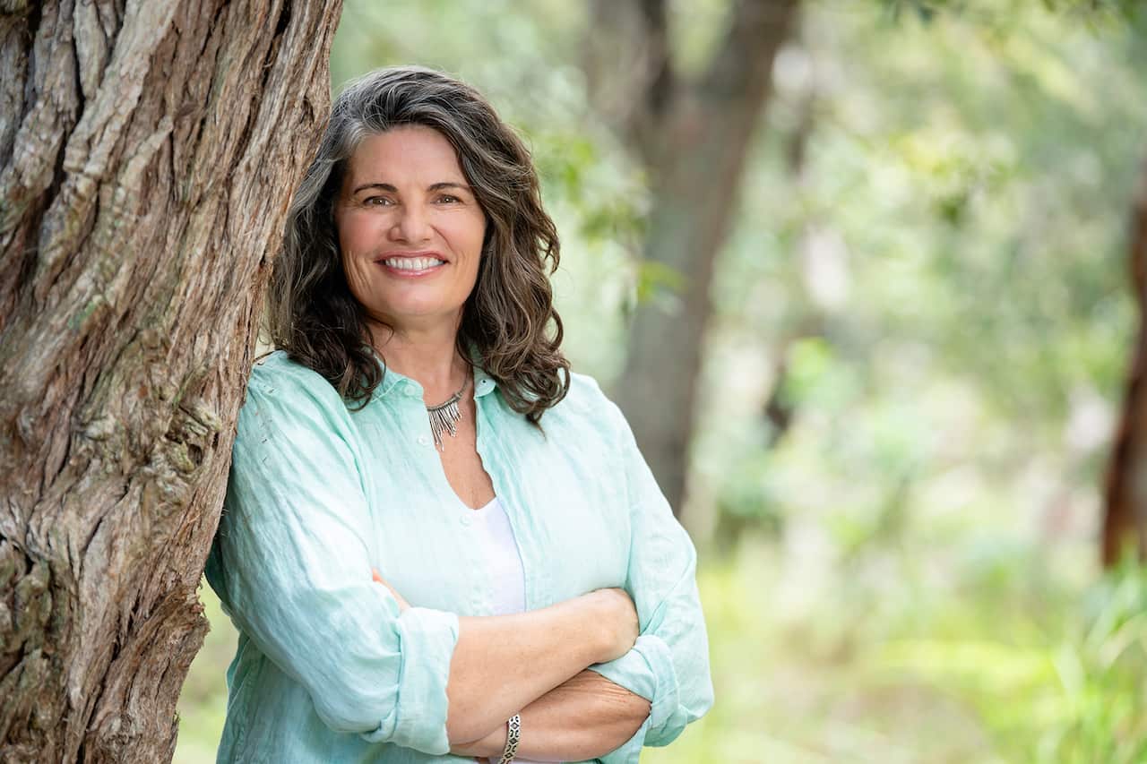 A smiling woman in a plae mint green shirt stands in the bush, leaning on the trunk of a tree 