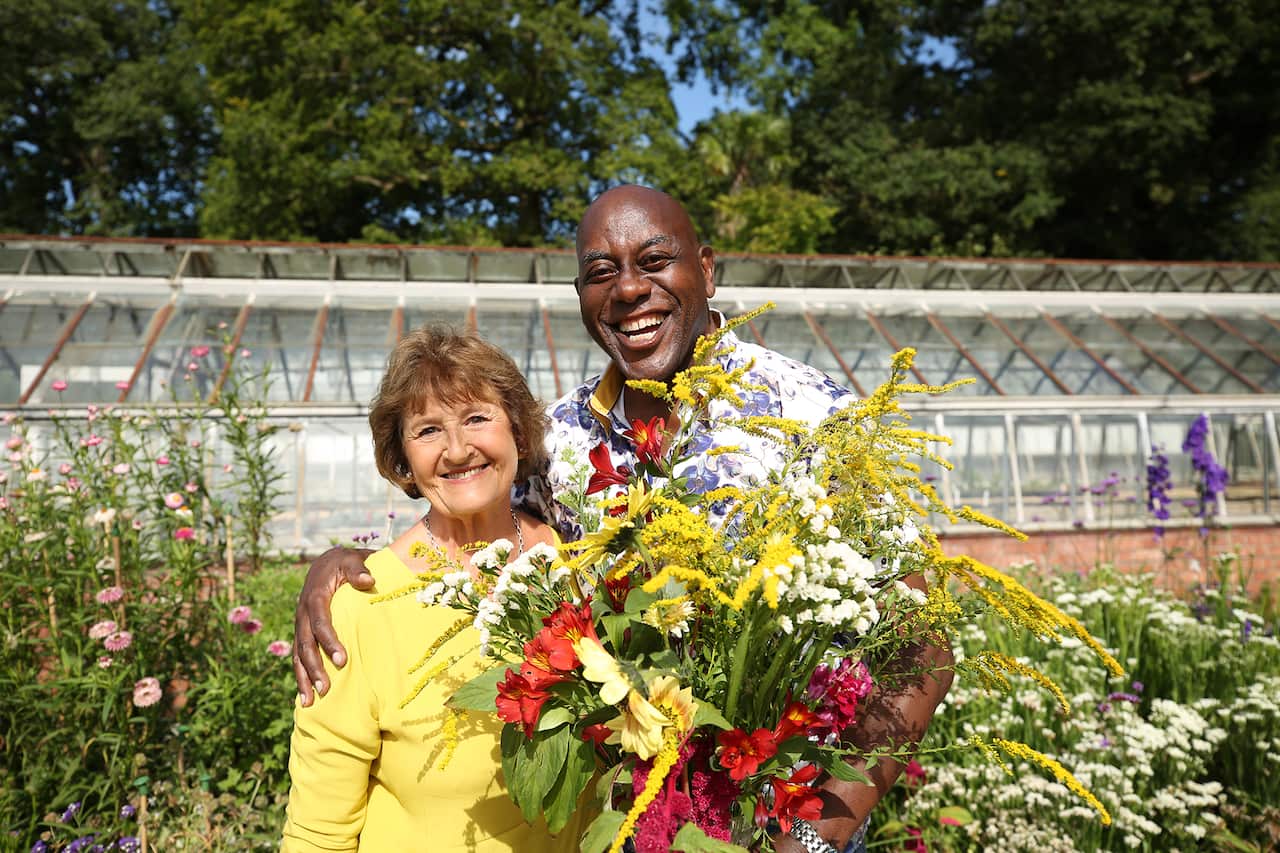Ainsley Harriott and volunteer stand in a flower garden. He is holding a big bunch of flowers. 