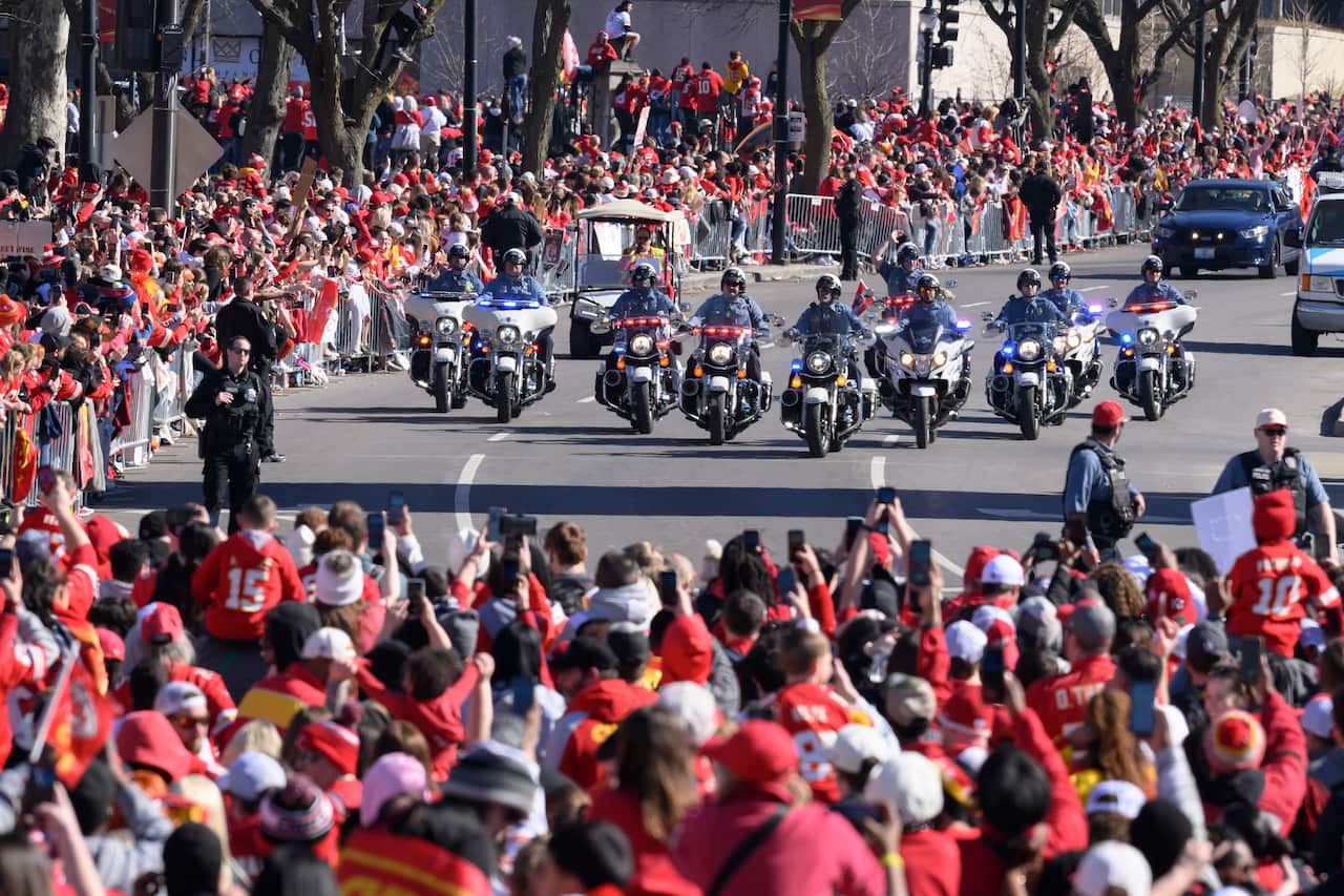 A police motorcycle motorcade rides between large crowds of people in red jerseys.