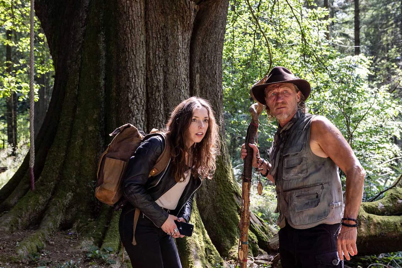 Scene in a forest, with the trunk of a large tree making up much of the background: A woman in a leather jacket, with a backpack, stands holding a phone, leaning slightly forward as if peering at something. A man with a long walking stick stands beside her, turning to look in the same direction.