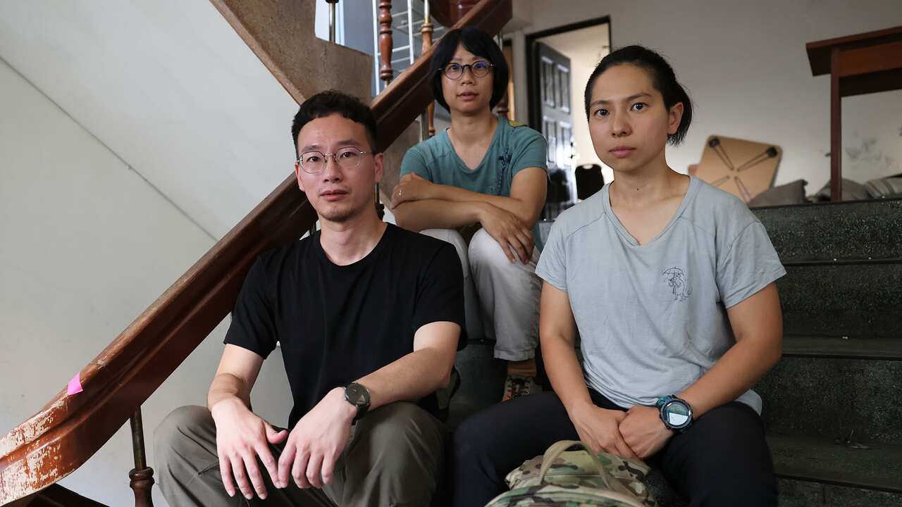 A young man wearing glasses and black t-shirt, a woman in grey t-shirt, and a woman wearing glasses and sage green t-shirt behind them are sitting on the stairs and looking into the camera.