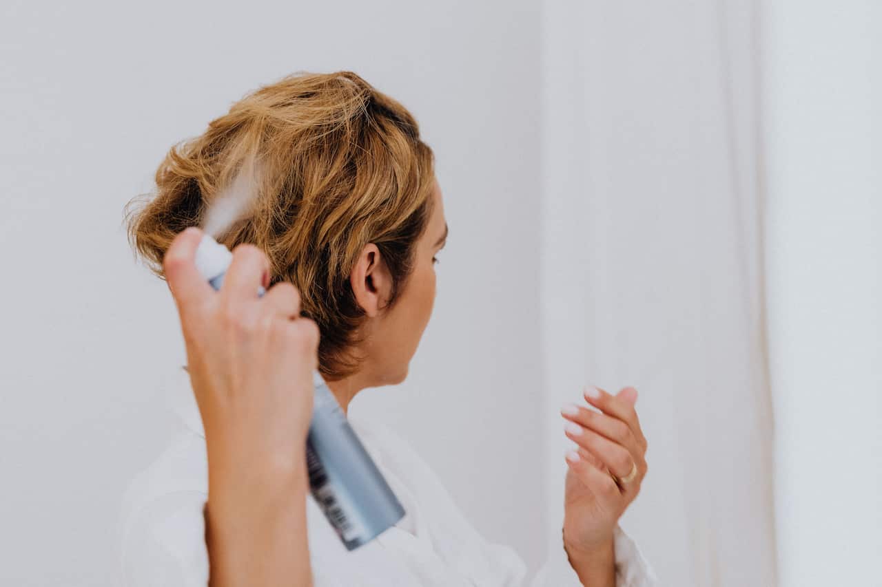 A woman in a white bathrobe sprays an aerosol product into her short, blonde hair.