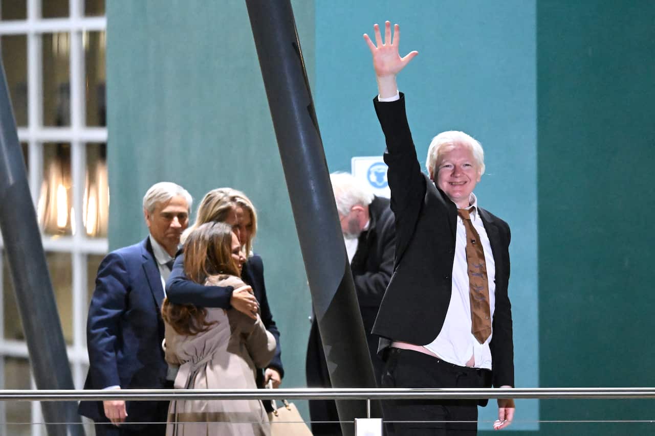 A man in a black suit and white shirt (Julian Assange) raises one arm above his head