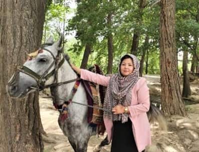 A woman in a pink cardigan and head scarf stands next to a grey horse.