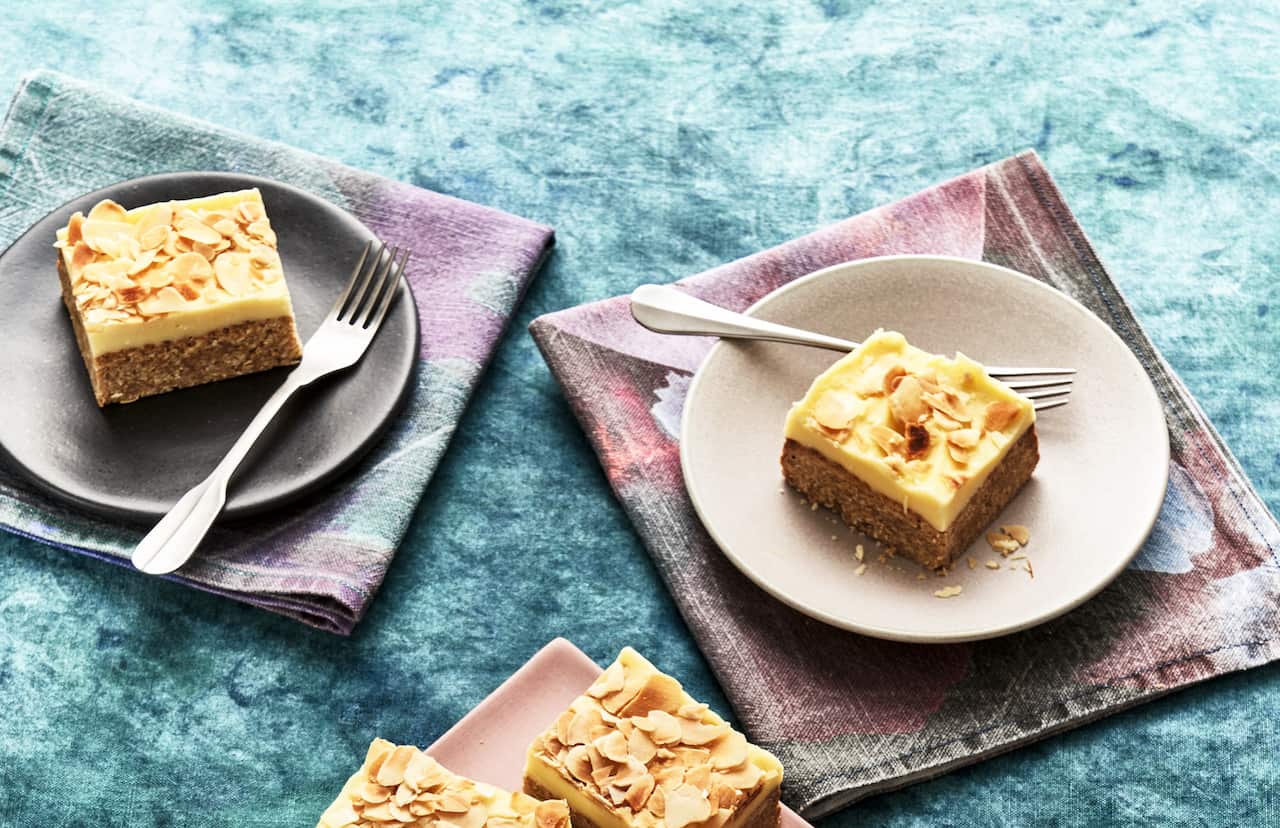 Deep squarish pieces of a layered bake, with a thick brownish bottom later and creamy top layer, scattered with flaked almonds, sit on wide plates. Each plate sits on a napkin in tones of blue, grey and pink. 