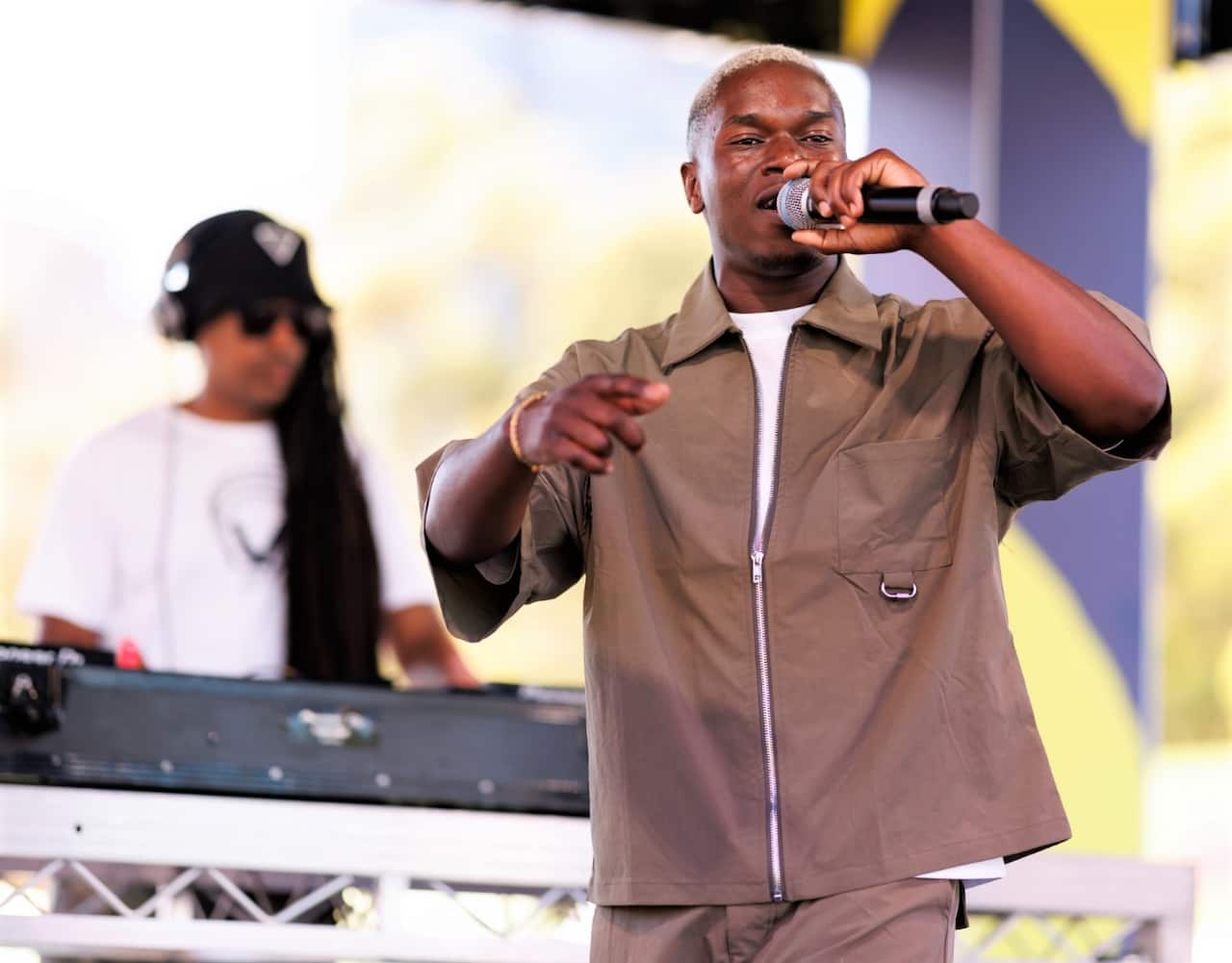 A man in a brown shirt stands holding a microphone in front of a mixing desk.