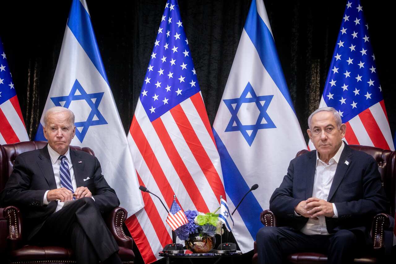 Two men wearing suits and ties seated in chairs. Behind them are the flags of the United States and Israel.