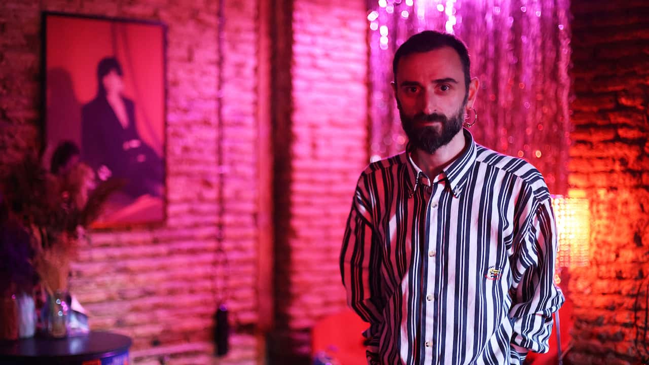 A man with a beard wearing a black-and-white striped shirt standing in a pink-lit bar.