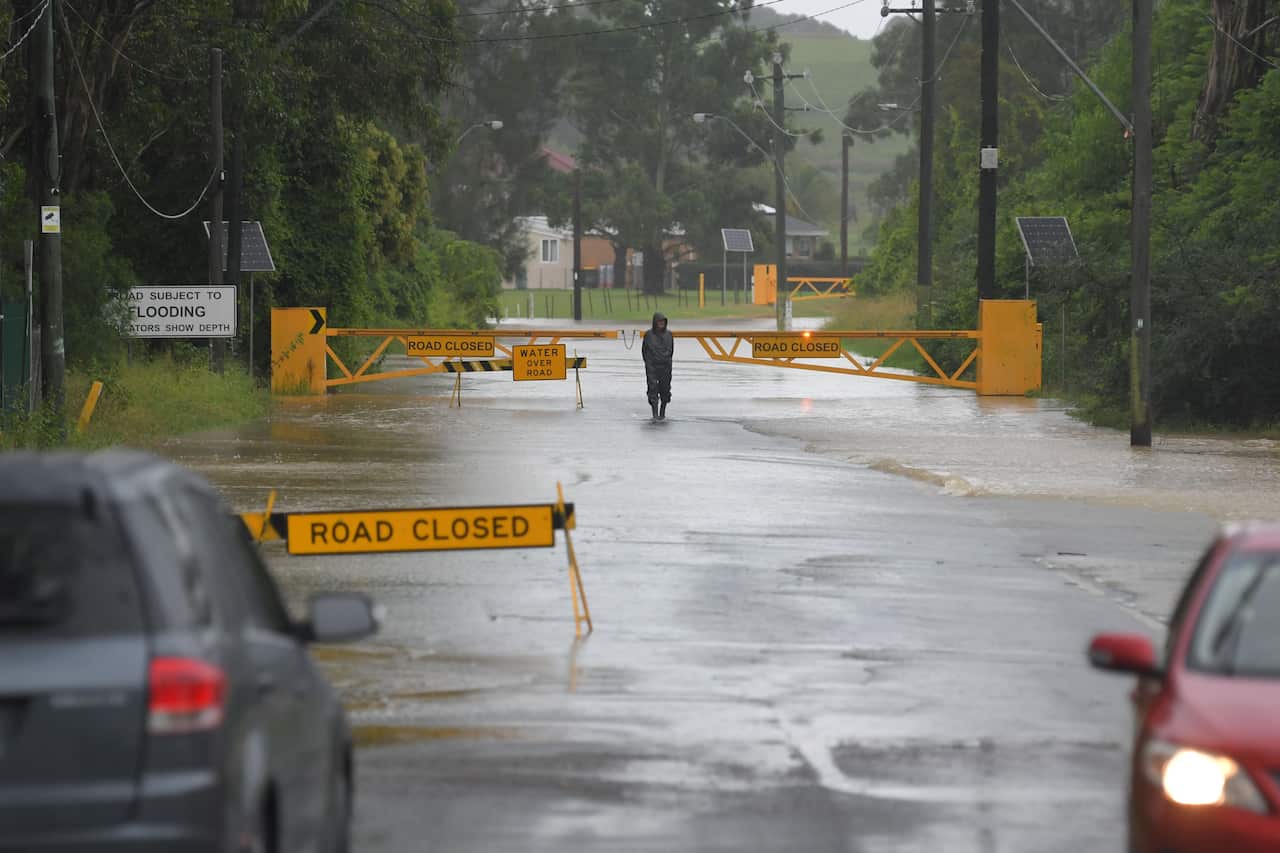 A man walks along a road flooded with water, with two cars and a yellow 'road closed' sign in view.