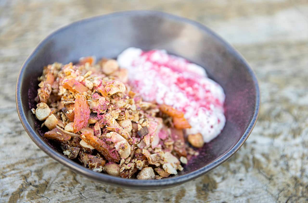 A dark bowl holds crunchy granola and yoghurt with a pink topping. 